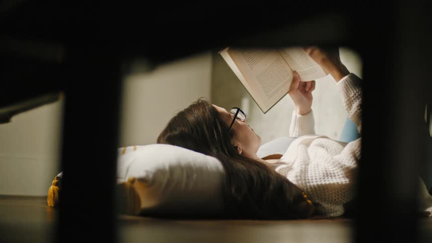 Young woman wearing glasses reads a book while laying on the bedroom floor
