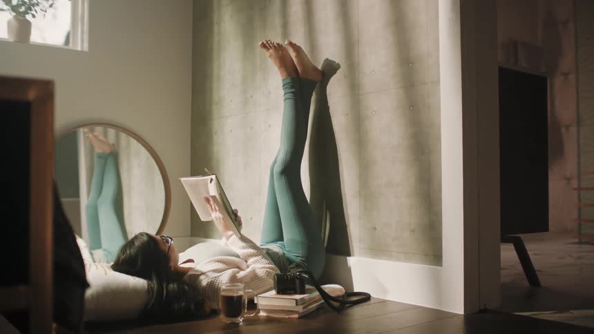 A woman is lying on her bedroom floor holding up a book as she reads with her legs up against the wall