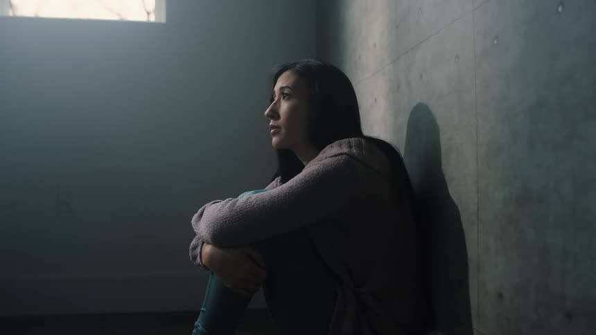 A woman is sitting on the floor leaning against the wall with her arms around her legs