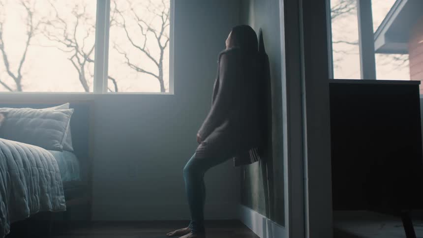 A woman walks into her bedroom to the wall and sinks to the floor with her hands on her head as she cries
