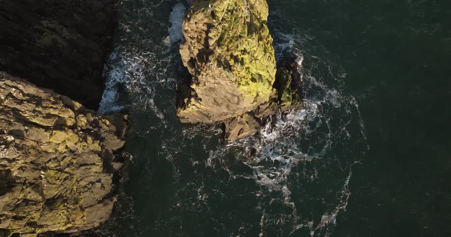 The sun shines upon tall rock formations with moss growing on the sides as waves splash against them