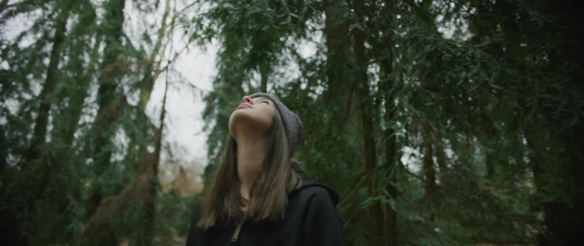 A teenage girl with long hair wearing a knit hat is standing in the woods looking up at trees
