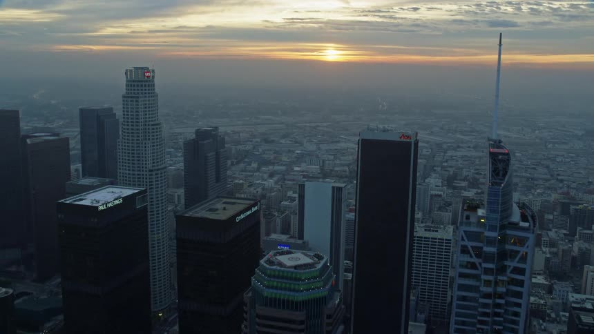 An epic morning flyover of Downtown Los Angeles skyscrapers as the sun rises behind them revealing Boyle Heights and warehouses along the Los Angeles River
