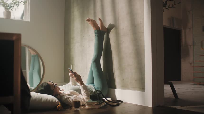 A woman is lying on her bedroom floor with her head on a pillow and her legs up against the wall as she texts on her phone