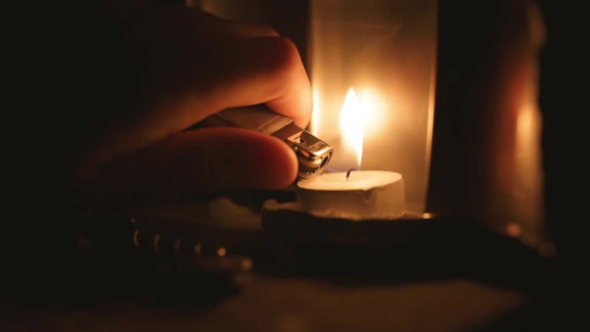 A persons hand ignites a lighter and lights a small candle as the flame glows in the darkness