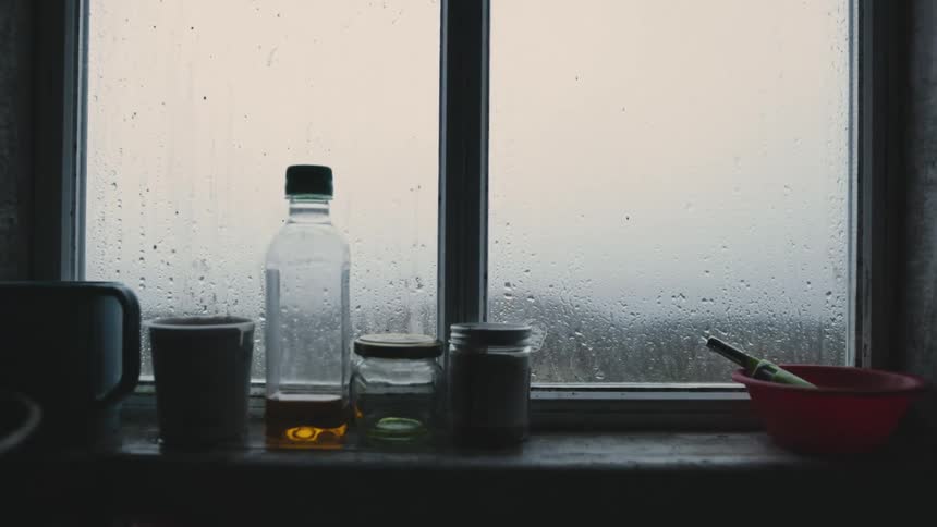 Bottles and dishes sit on windowsill as raindrops run down glass