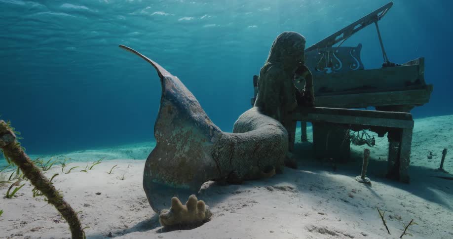 A statue of a mermaid and an old piano sit underwater on the sea bed as light shines from above