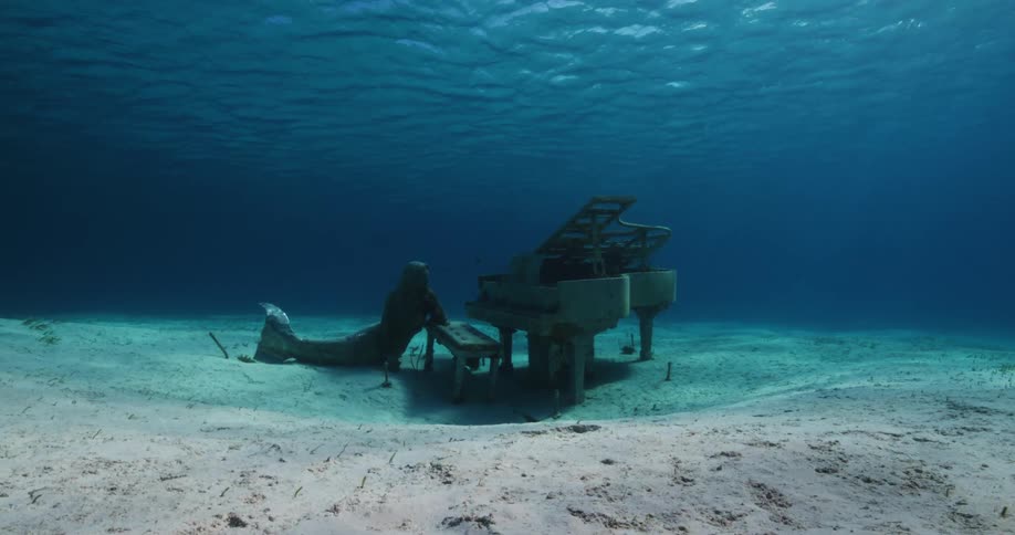 Statue of mermaid leaning against piano bench near piano sits on seafloor