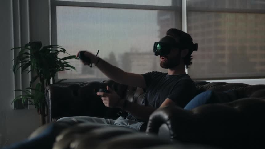 A man wearing a vr headset sitting on his couch moving his hands out and around in front of him