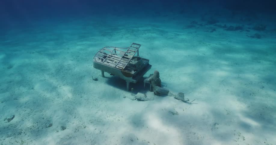 An old piano and a statue sit underwater on the sea bed as light shines from above