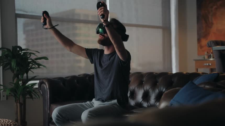 A man wearing a vr headset sitting on his couch raising his arms and talking