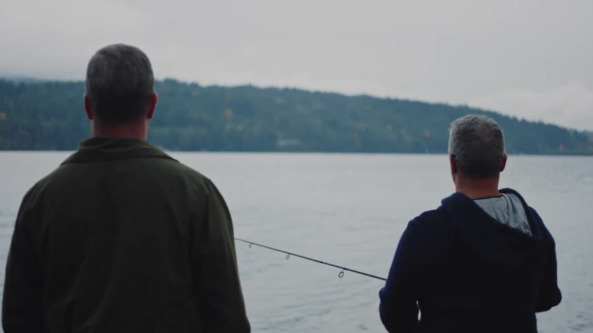 One man stands next to another who is fishing with a rod and reel across water from forested hill