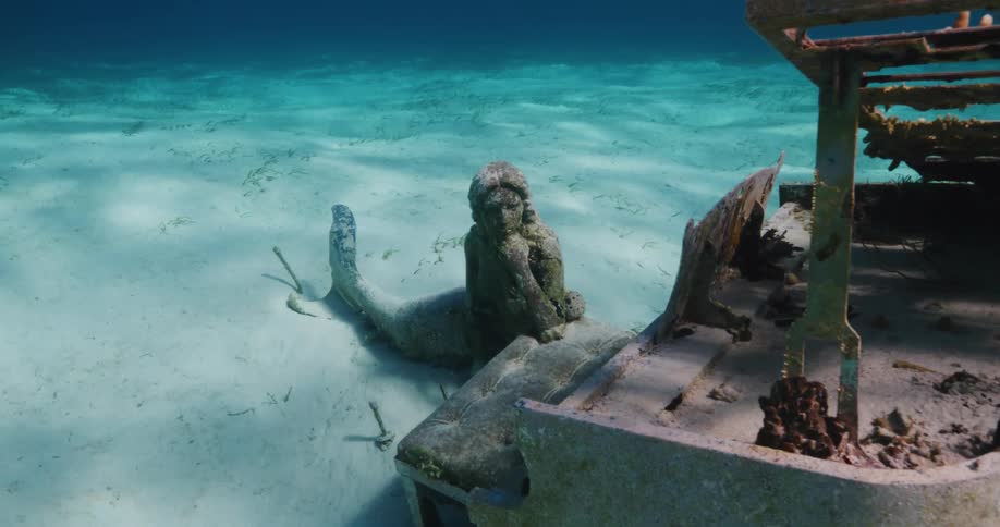 A statue of a mermaid sits among shipwrecked items in the ocean