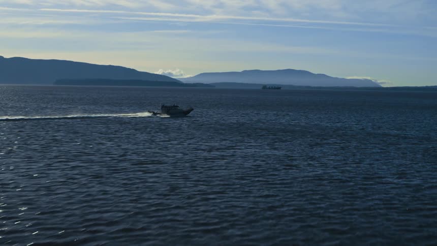 A light flashes on top of a boat as it sails across the water and there are mountains in the distance