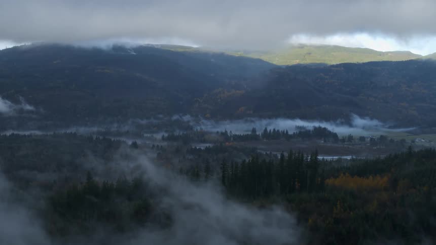 Dark clouds move over a mountain river valley as fog rises among the trees and there are mountains in the distance