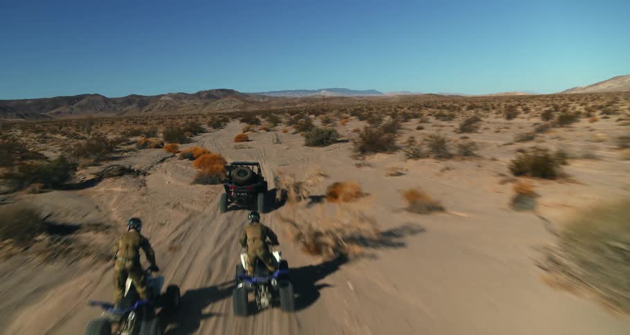 Two people on four wheelers follow a jeep across arid scrub land toward mountains