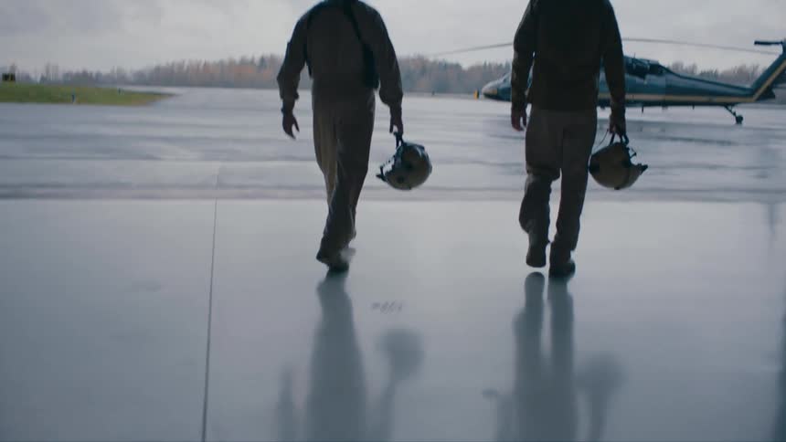 Two pilots carrying helmets walk out of hanger to parked helicopter