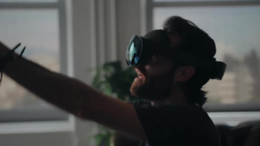 A close up of a man wearing a vr headset sitting raising his arms and hands and talking