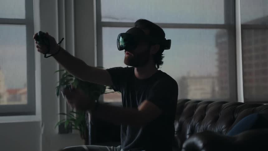 Man sitting on couch near window wears vr goggles and interacts using handheld controllers