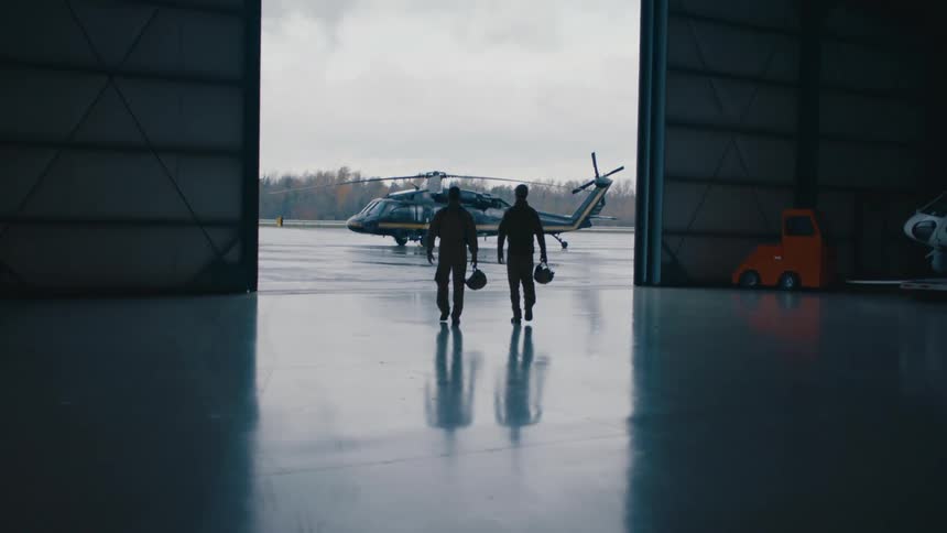 Two men carrying bags are walking out of a hangar and toward a helicopter
