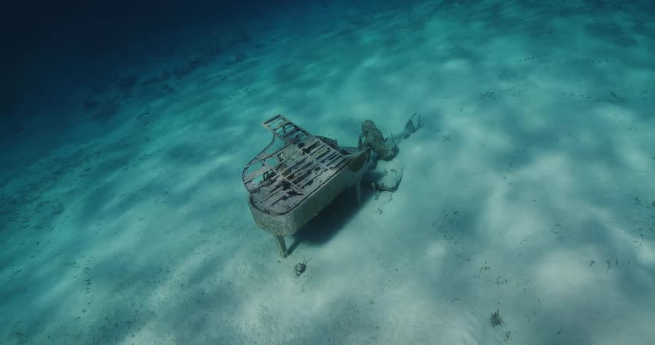The statue of a mermaid leaning against a piano under water on the ocean sandy bottom