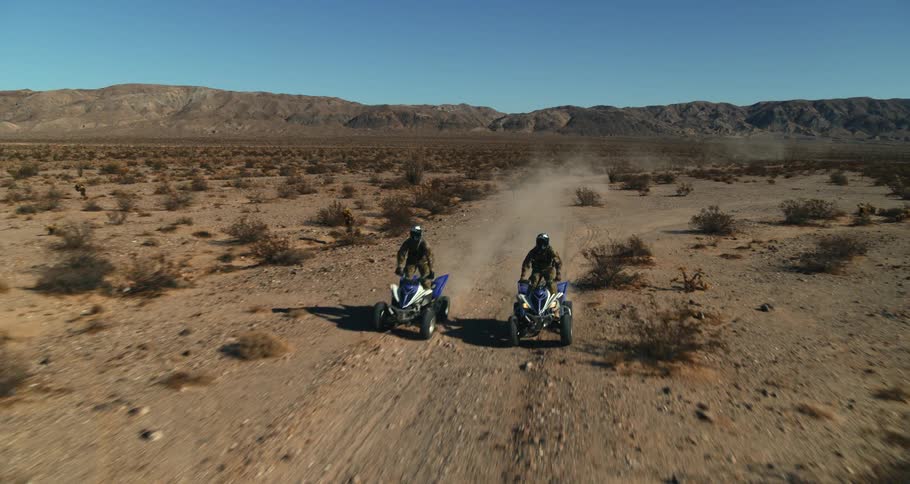 People riding four wheelers pass intersection of two dirt roads in arid scrub land