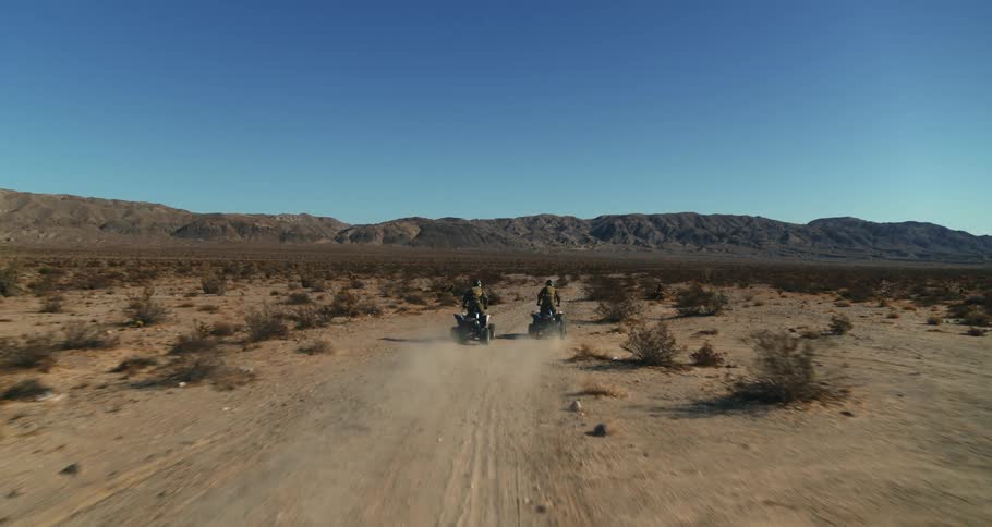 Two people ride four wheelers along dirt path through arid scrub land toward mountains under a clear blue sky