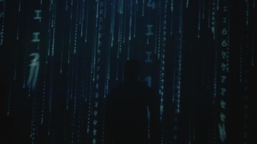A man walks through a dark room as binary code is flashed as a projection