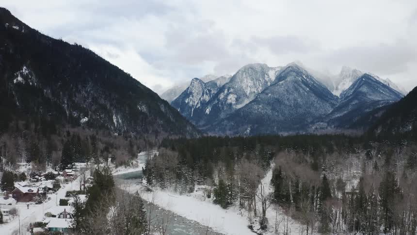 An aerial of a small town at the bottom of a mountain range covered in snow next to a river and a forest