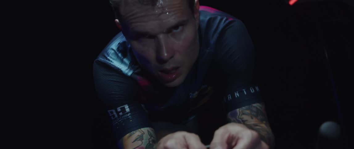 Sweaty tired man with tattoos pedals stationary bike in dark room