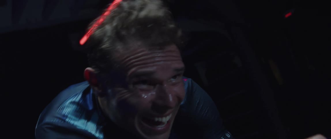 Twisting footage of a man pedaling on a static bicycle inside a dimly lit room against a dark background