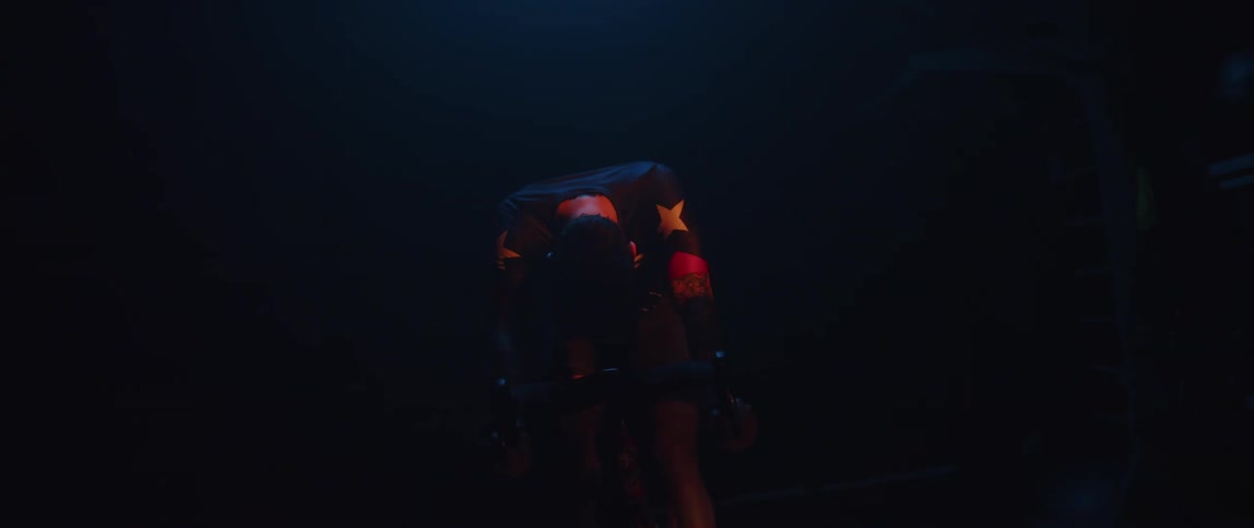 Man wearing a cycling jersey and cycling glasses lifting his head up while sitting on a stationary bike in a dark room