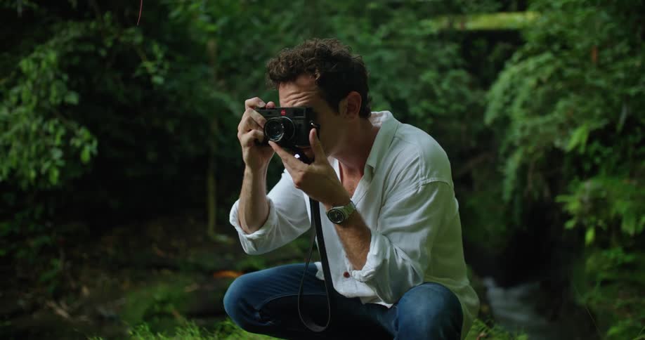 A man is sitting down among tropical plants holding his camera and looking around when he lifts his camera and takes a picture