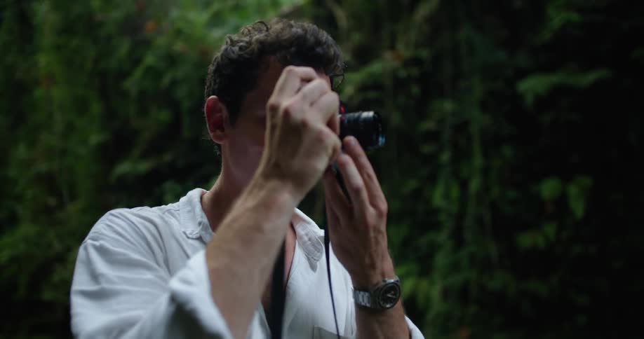 A man is standing by a stream holding his camera and taking a picture and then looking up at the trees