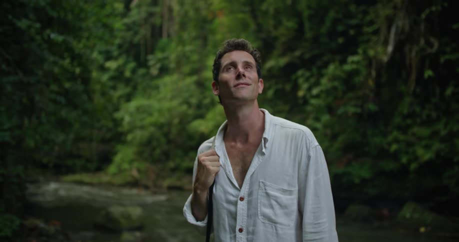 A man is standing by a stream holding his camera strap and looking up at the trees in the rainforest
