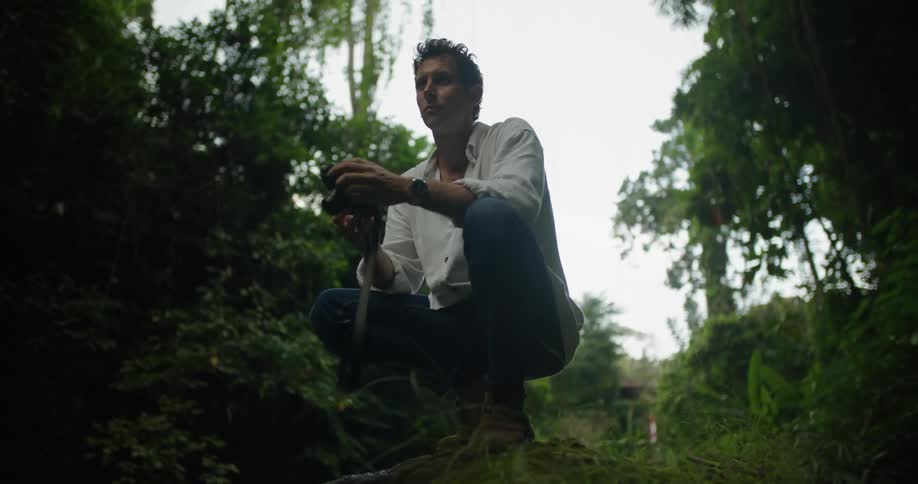 A man is squatting down on a rock in the rainforest holding his camera and taking a picture