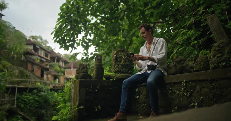 Man sitting on a wall decorated with small sculptures in the jungle uses his smartphone