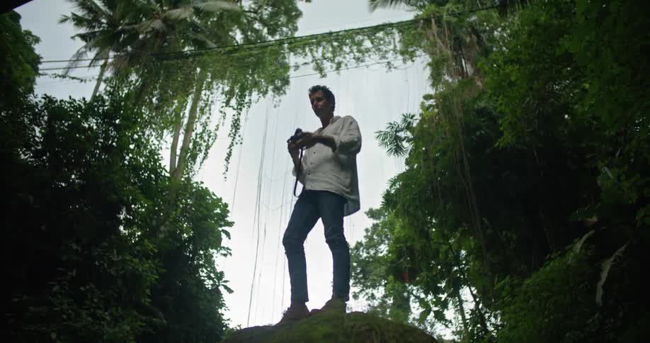 A man is standing on ledge surrounded by tropical plants and he takes the camera from his shoulder and takes a picture