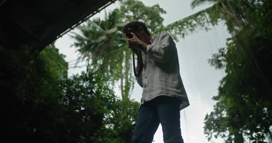 A man is standing on ledge surrounded by tropical plants and he takes the camera from his shoulder and takes a picture