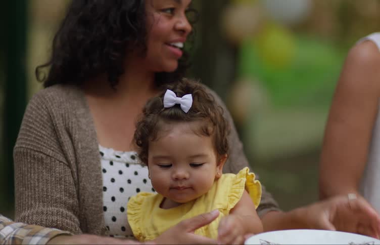 Mother smiles and holds daughter in her lap as she talks to others while sitting at table outdoors