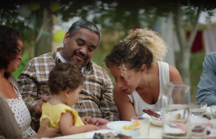A family sits at a table outdoors while they interact with a toddler in a yellow dress