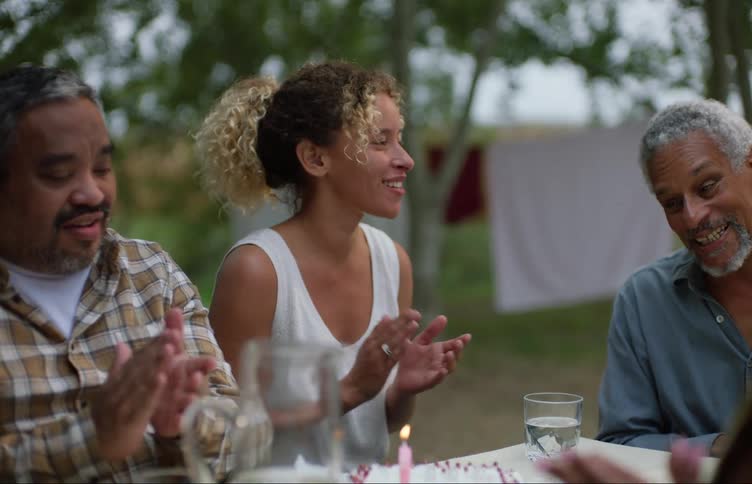 A woman brings a birthday cake with lit candles to a table as people sing to a man