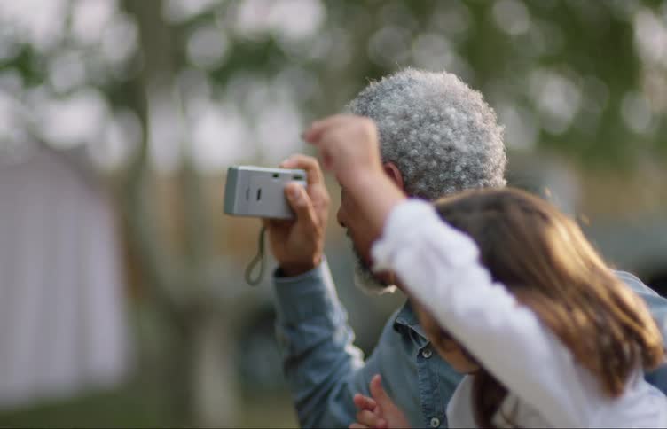 Grey haired man smiles and uses a camera as a young girl leans in from the right