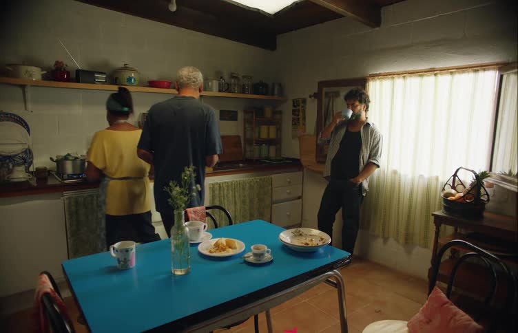 Man and woman converse by the sink as a man leans against the window drinking coffee