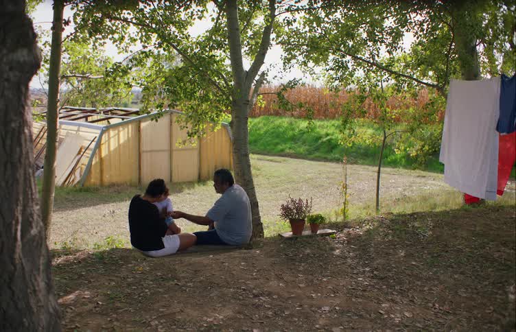 Grandfather and daughter sit with her child on ground overlooking barn with crops in the nearby field