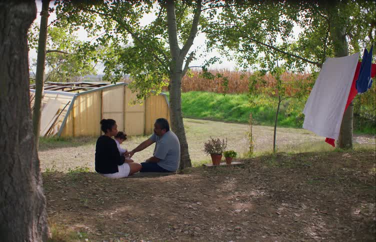 A man sits outside and puts his hand on his daughters arm as he talks with her while she holds her daughter