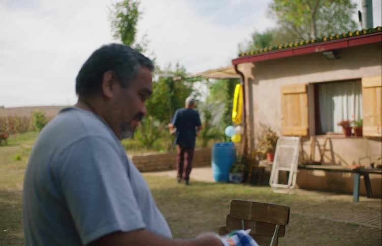 Man in blue shirt dries plate and looks at woman carrying table in front of building in the countryside