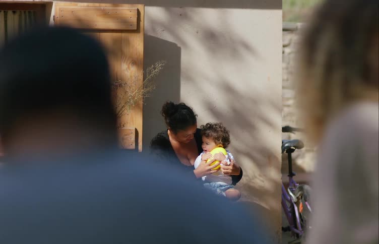 A woman plays with a baby sitting on her knee as others walk past