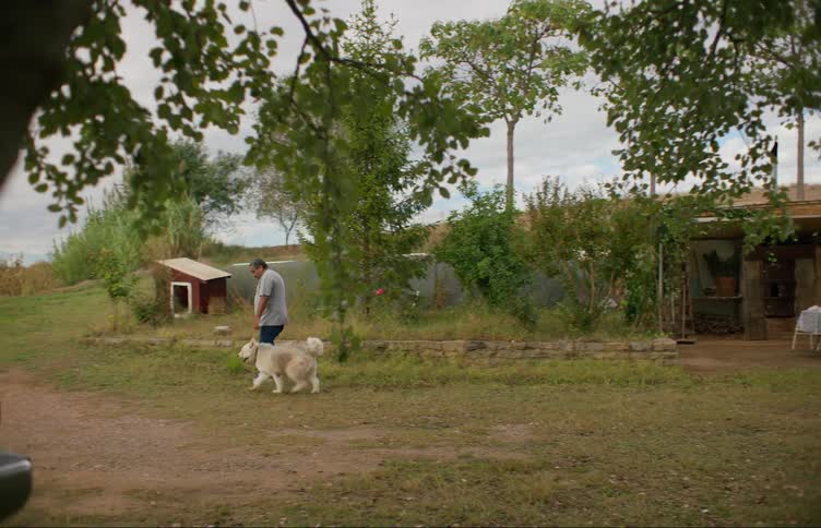 A man walks out of his home in a rural area with a dog and leads him to a dog house where he ties him up