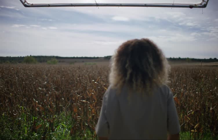A woman with very curly hair stands up and looks out upon a cornfield